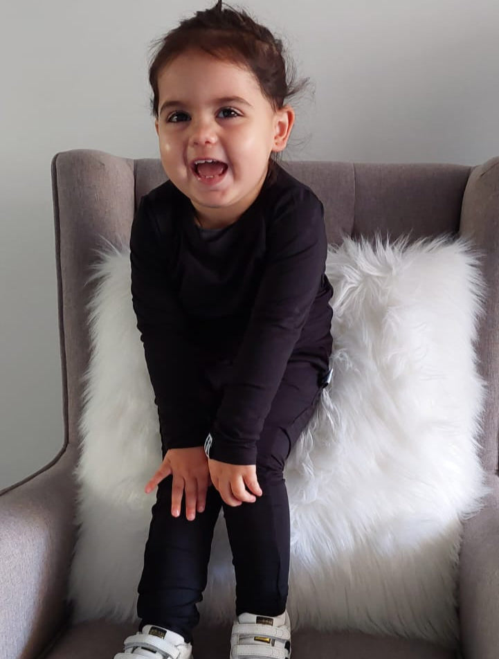 Child sitting on a gray chair with a white fur pillow, wearing a dark outfit and white sneakers.