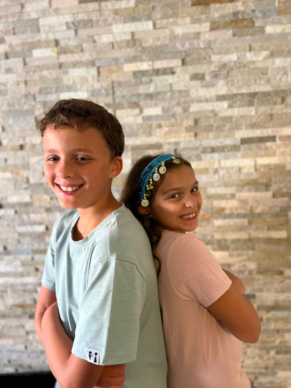 Two children standing back-to-back in front of a stacked stone wall. The child on the left is wearing a light sage green t-shirt, and the child on the right is wearing a light pink t-shirt and a blue beaded headband.
