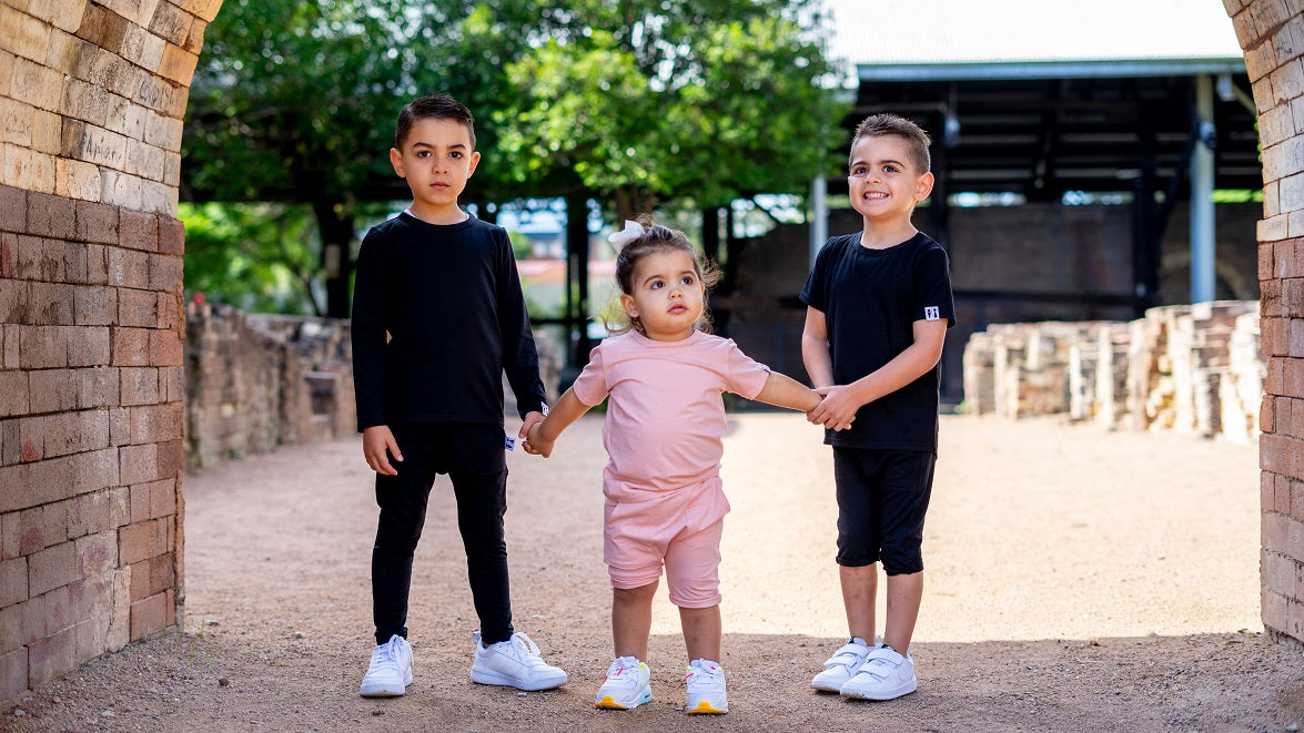 Three children standing hand-in-hand in a brick archway, wearing matching black and pink outfits.