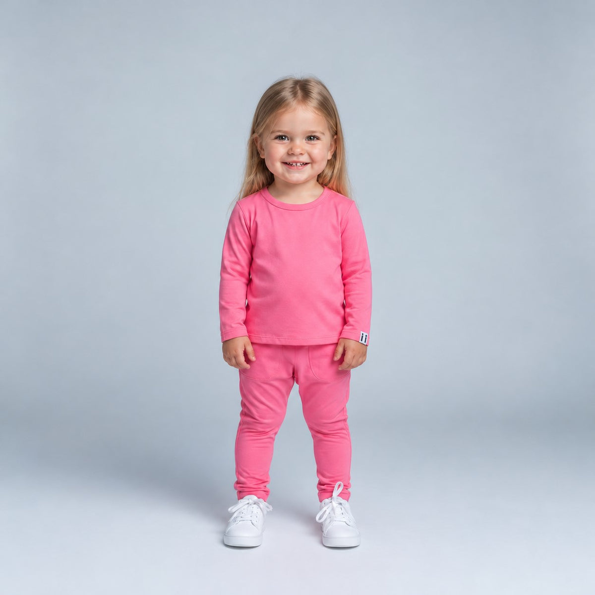 A young girl with blonde hair stands smiling, wearing a matching bright pink long-sleeve top and jogger pants set, paired with white sneakers, against a light gray studio background.