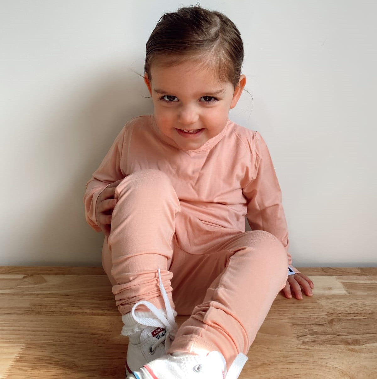 A young child wearing a matching dusty pink long-sleeve top and jogger pants set, sitting on a wooden floor against a plain light wall.