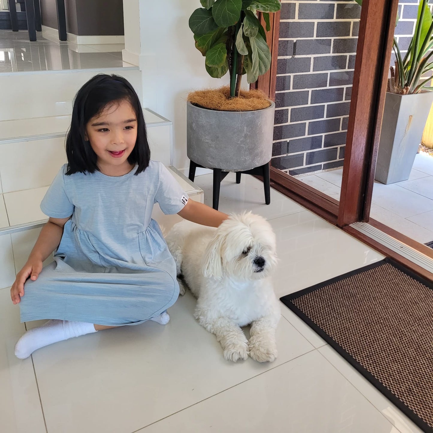 Young girl in a blue dress sitting on the floor with a white dog, surrounded by indoor plants.