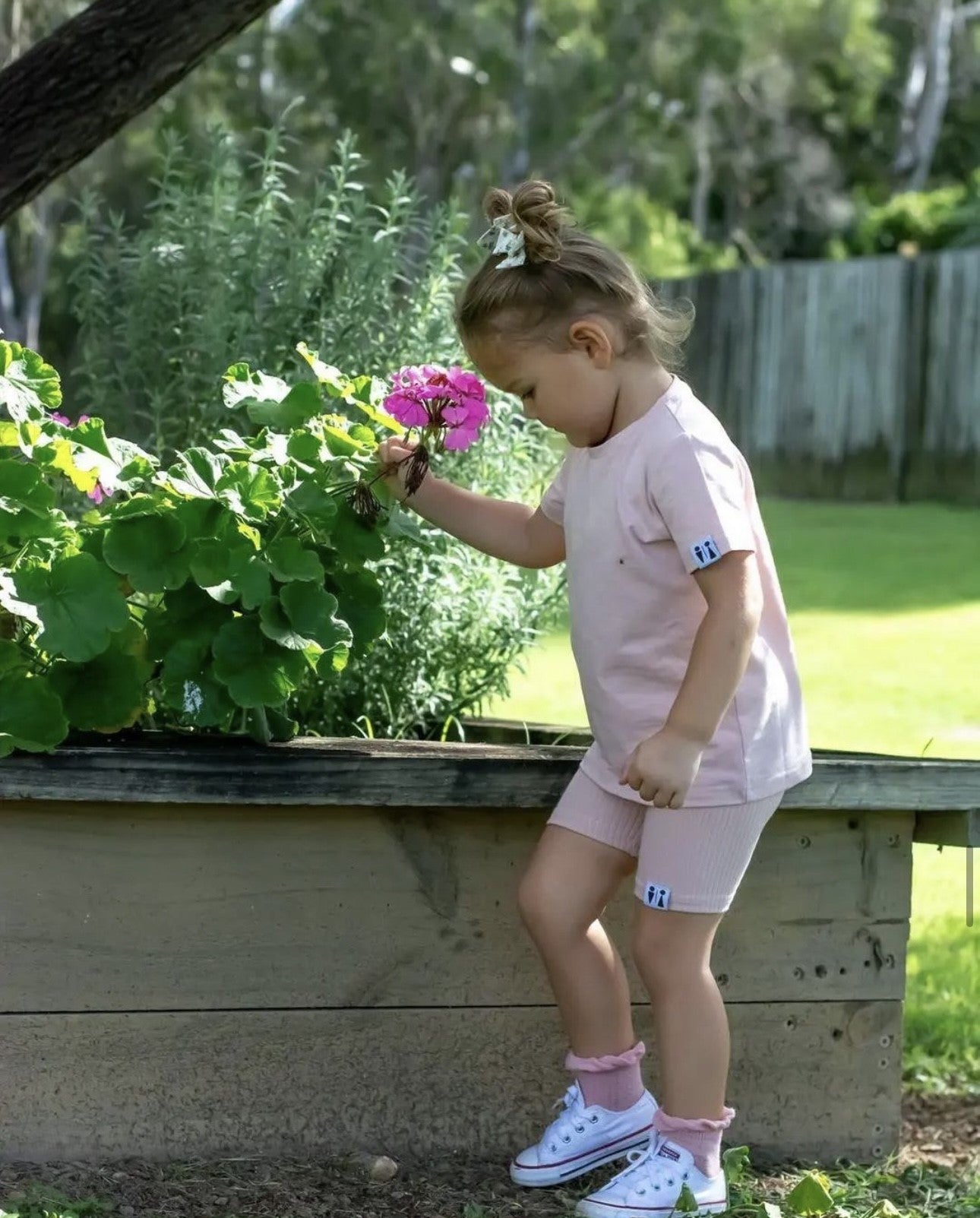 A young girl in a light pink t-shirt and matching shorts stands in a garden next to a wooden raised bed, touching a bright pink flower. She is wearing white sneakers with pink socks.