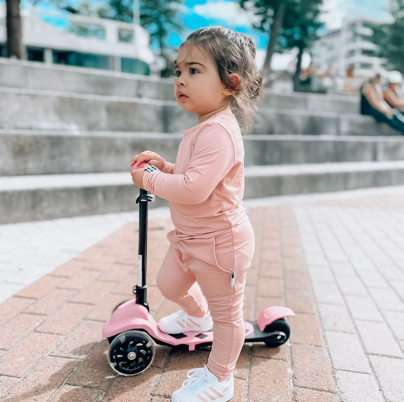 Child in pink bamboo Baby Todd & Co. outfit standing next to a pink scooter on a sidewalk.