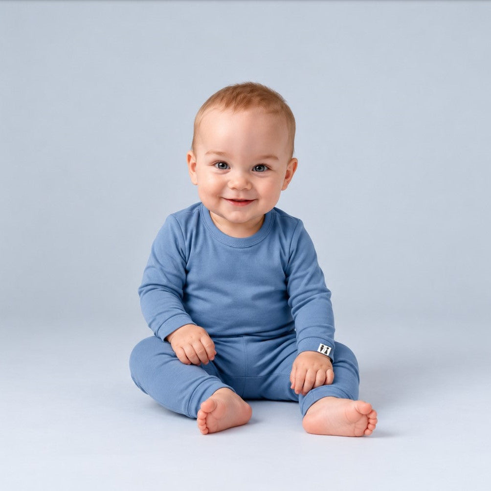 A baby sitting cross-legged wearing a solid medium blue long-sleeve one-piece pajama or romper, photographed against a seamless light gray studio background.